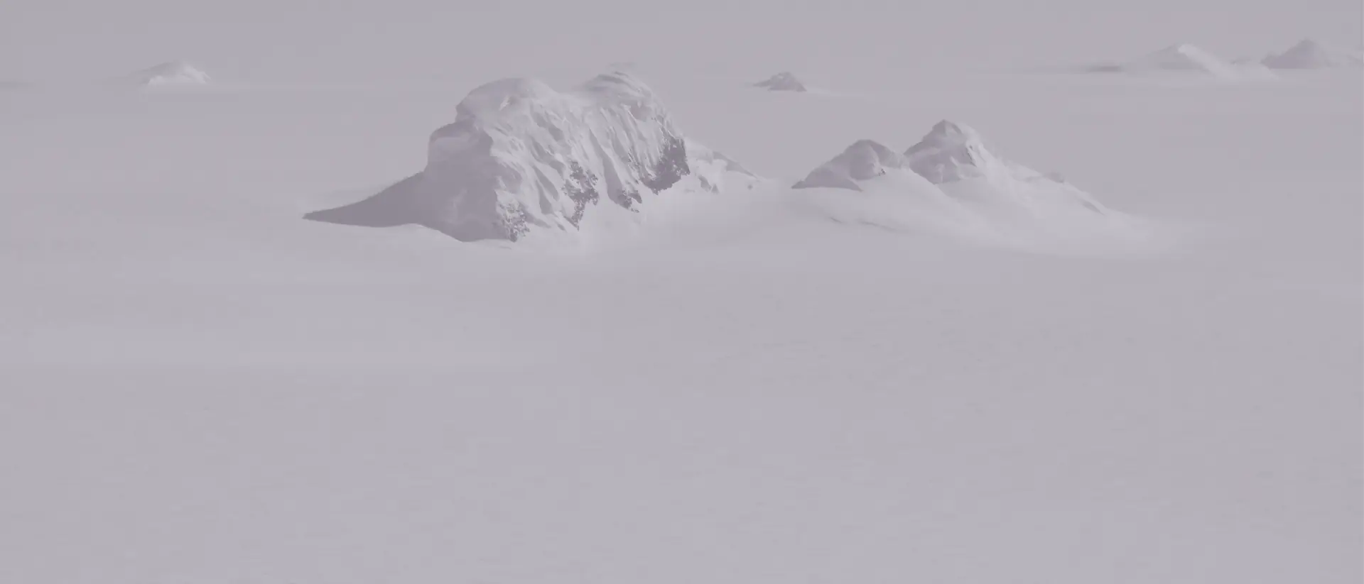 Luftaufnahme der Antarktis: einzelne Felsgipfel ragen aus einer weiten, flachen Eisdecke, darüber ein blasser Himmel